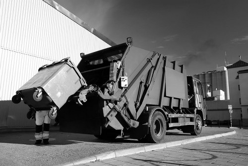 An industrial rubbish collection vehicle, fitted with a hydraulic lifting arm, is in the process of emptying a large wheeled skip container filled with mixed waste, including cardboard boxes and plastic debris. The skip, made of metal with a textured surface, is positioned on a paved urban road adjacent to a tall corrugated metal fence, which appears to enclose a commercial or industrial site. The vehicle’s cab is visible, and the truck is equipped with safety features such as rearview mirrors. Behind the vehicle, there are various warehouse buildings and infrastructure typical of an urban environment, with the sky overhead partly cloudy. The scene illustrates a professional rubbish removal operation, possibly carried out by Office Clearance Shepherds Bush, in a residential or commercial area within London or nearby, as suggested by referencing Shepherds Bush. The overall setting emphasizes efficiency in waste collection in a city context, supporting rubbish clearance and disposal services.