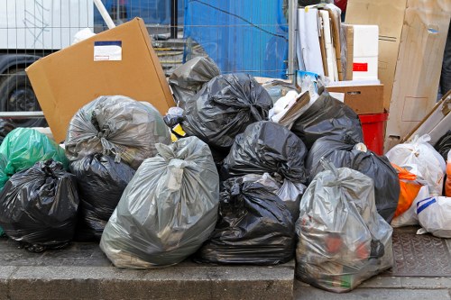 A collection of black and grey plastic rubbish bags, filled with various waste materials, are piled on a concrete pavement outside, possibly in an urban or residential area within Shepherds Bush. Some bags are tightly knotted, while others appear loosely tied, with contents not fully visible but suggesting mixed household or office waste. The bags are stacked irregularly, with a few resting against each other and some leaning to the side. In the background, there are cardboard boxes, a blue tarpaulin or covering, and other miscellaneous packaging materials, indicating a clearance or disposal process. The scene is outdoors, under natural light, with nearby building and street elements hinting at a typical rubbish collection situation suitable for a service like Office Clearance Shepherds Bush, specialising in waste removal and rubbish clearance in the local area of West London.