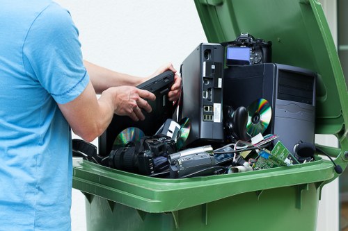 A person wearing a blue shirt is seen placing electronic waste, including old computer towers, DVDs, and various small electronic devices, into a green rubbish bin outdoors. The contents include a black desktop computer tower with a partially visible side panel, several compact discs, and miscellaneous electronic components, all arranged on the bin's open lid. The environment appears to be a driveway or paved area, with the focus on the electronic waste being prepared for disposal or collection. The scene reflects a professional rubbish removal process, consistent with services provided by companies like Office Clearance Shepherds Bush, in an area near Shepherds Bush. The lighting is natural, and the electronic items are stacked loosely, with some items leaning against each other, demonstrating the typical approach to disposing of electronic waste responsibly.