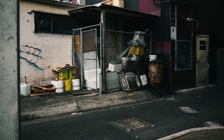 Four black wheelie bins with closed lids, each marked with the number 33, are positioned on a paved sidewalk in front of a brick building. The building's exterior wall features a combination of painte