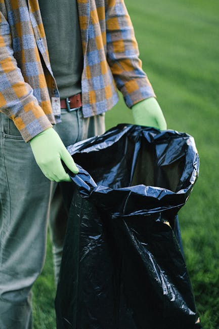 A person dressed in orange work overalls and white footwear is holding two large blue plastic rubbish bags, each filled with waste material, one in each hand. The bags appear semi-transparent, reveali