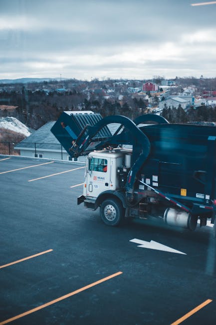 A waste collection vehicle parked on a narrow cobblestone street adjacent to multi-storey residential buildings with weathered facades and peeling paint. The rear of the white garbage truck is open, r