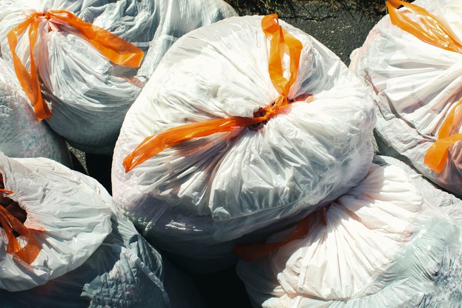A person's hand and forearm are visible, grasping a green plastic bag with blue and white elements, filled with waste material. The bag is tied at the top with a twisted section of plastic. The backgr