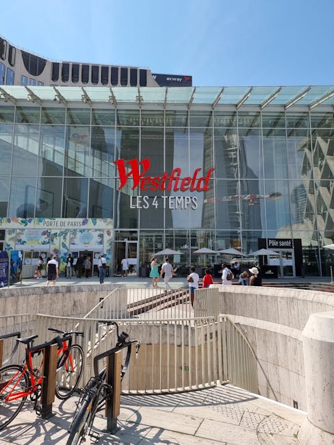 The image depicts the front entrance of Westfield London shopping centre with a large glass facade reflecting surrounding buildings and a clear blue sky. The centre's prominent red and white signage r